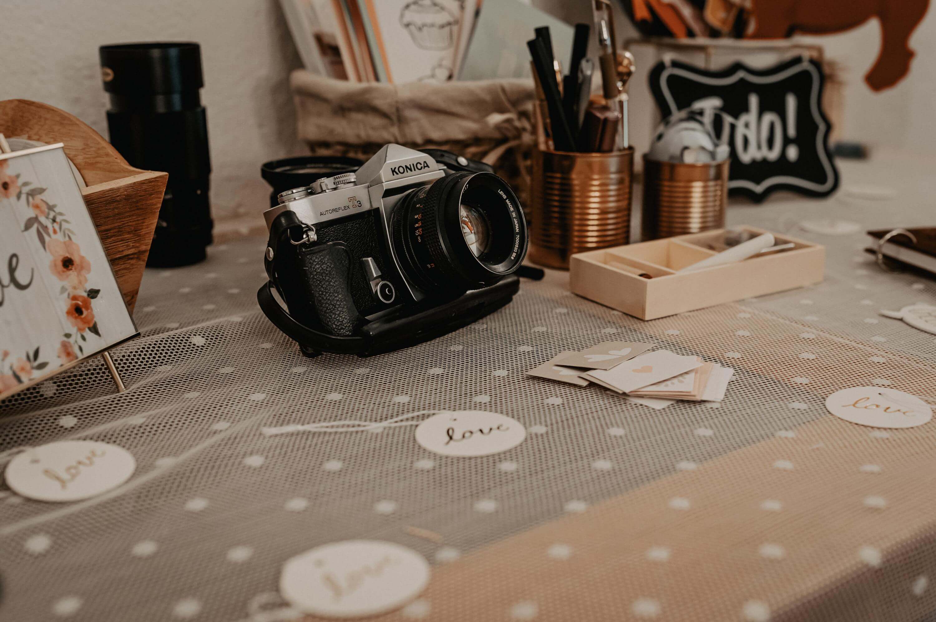 Hochzeit Feier Burg Kleeburg und Boho Auf einem Sideboard mit weißem Tischtuch und kleinen runden Papierbadges liegt ein alter analoger Fotoapparat neben Dekorationsartikeln wie Kupferblechdosen mit Schreib-Utensilien für das Gästebuch einer Hochzeitsfeier.